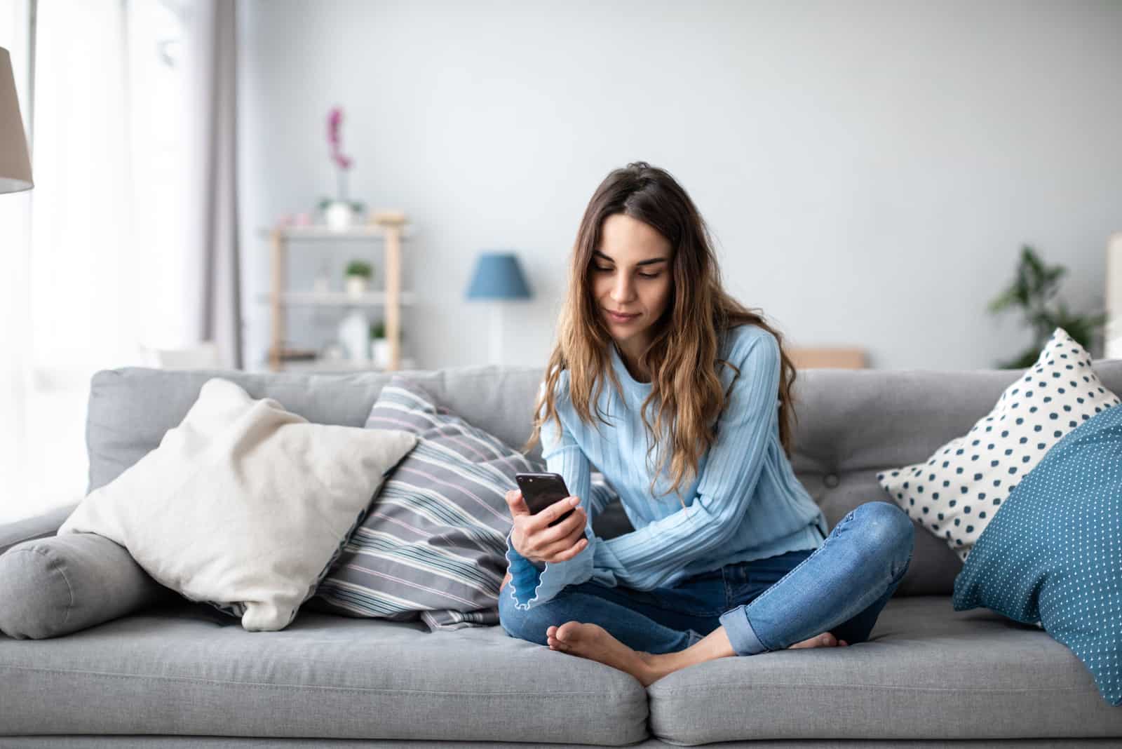 Une femme aux longs cheveux noirs est assise sur le canapé avec un téléphone à la main