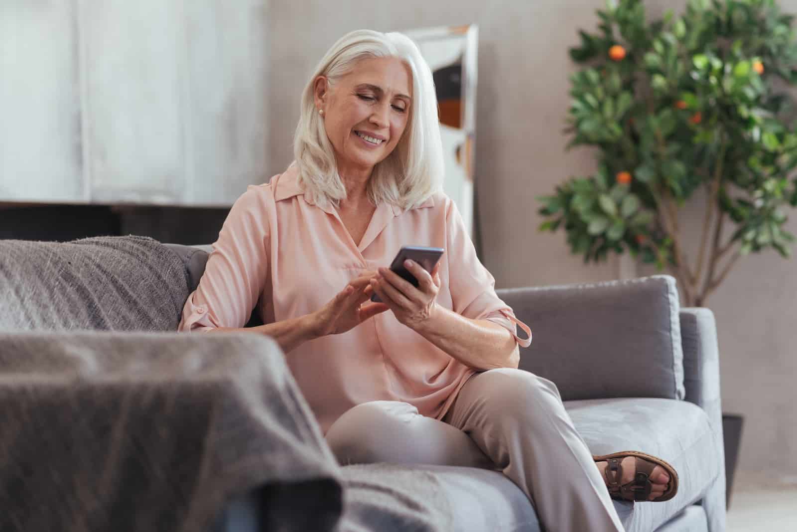 une femme plus âgée souriante assise sur le canapé et tapant au téléphone